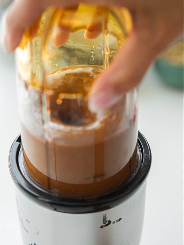 A close-up of a hand holding a small blender with a brown liquid being blended inside a transparent container. The blender base is white and black, and the background is blurred.